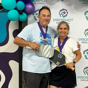 Couple holding pickleball paddles celebrate a win, wearing medals at a ProShot Pickleball event, smiling for photo.